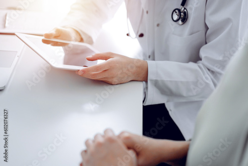 Doctor and patient sitting at the desk in clinic office. The focus is on female physician's hands using tablet computer, close up. Perfect medical service and medicine concept