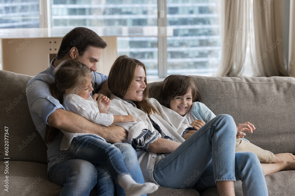 Cheerful young parents playing with little children on couch at home, enjoying family leisure, cuddling, tickling kids, giggling, laughing, having fun, feeling happiness, bonding, love Cheerful young parents playing with little children on couch at home, enjoying family leisure, cuddling, tickling kids, giggling, laughing, having fun, feeling happiness, bonding, love