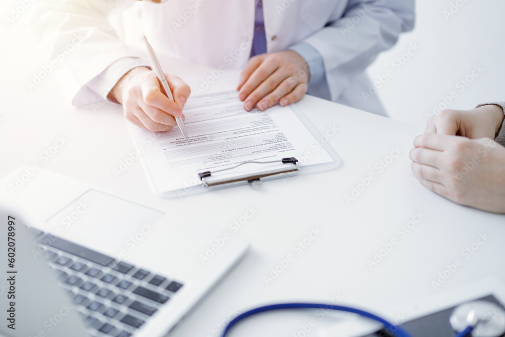 Doctor and patient sitting at the table in clinic. The focus is on female physician's hands filling up the medication history record form or checklist, close up. Medicine concept Doctor and patient sitting at the table in clinic. The focus is on female physician's hands filling up the medication history record form or checklist, close up. Medicine concept