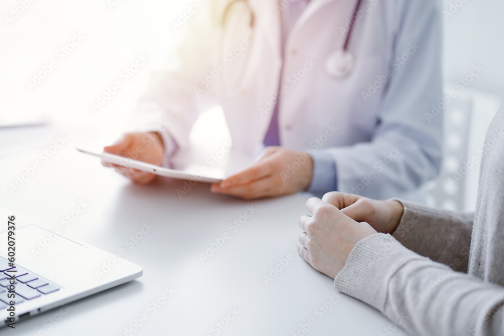 Doctor and patient sitting at the table in clinic. The focus is on female physician's hands using tablet computer, close up. Medicine and healthcare concept Doctor and patient sitting at the table in clinic. The focus is on female physician's hands using tablet computer, close up. Medicine and healthcare concept