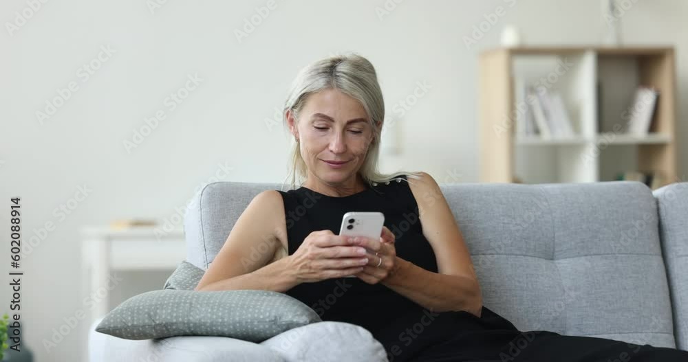 Cheerful mature woman typing text message on mobile phone, resting on home sofa, smiling, laughing, looking away, thinking dreaming, enjoying online communication, Internet service, app work Cheerful mature woman typing text message on mobile phone, resting on home sofa, smiling, laughing, looking away, thinking dreaming, enjoying online communication, Internet service, app work