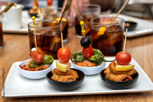 Tapa de pinchos típicos de la gastronomía española, acompañados de unos vasos con vermut de fondo