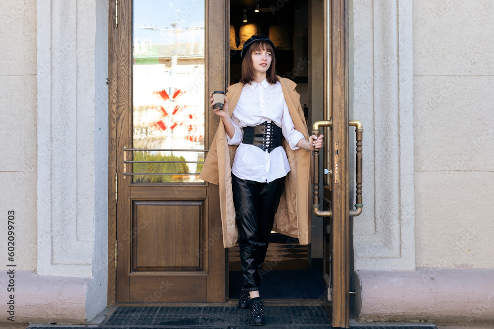 A lovely young brunette with a bob haircut, wearing a beret and a beige classic coat, steps out of the coffee shop, opens the door of the building. A lovely young brunette with a bob haircut, wearing a beret and a beige classic coat, steps out of the coffee shop, opens the door of the building.