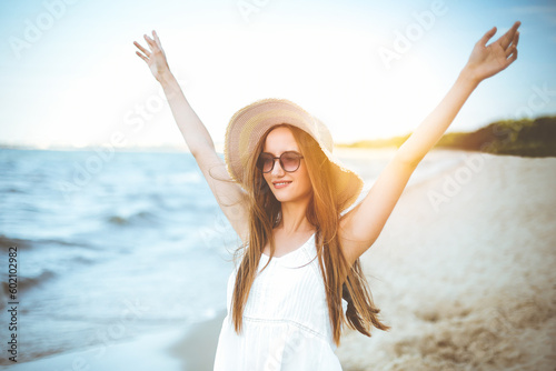 Portrait of a happy smiling woman in free happiness bliss on ocean beach standing with a hat and sunglasses. A female model in a white summer dress enjoying nature during travel holidays vacation