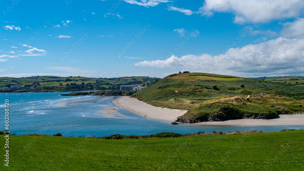 The green hills of Inchydoney Island and the famous Irish beach on a sunny summer. Clear blue sky with white clouds over the Irish coast. Irish summer landscape. Green grass field near water