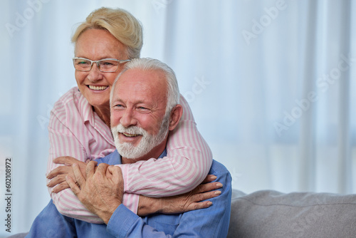 Happy senior classy couple hugging, bonding sitting at their house or nursing home. Cheerful elderly wife embracing husband looking away dreaming, enjoying wellbeing and love
