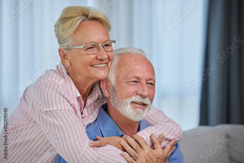 Happy senior classy couple hugging, bonding sitting at their house or nursing home. Cheerful elderly wife embracing husband looking away dreaming, enjoying wellbeing and love