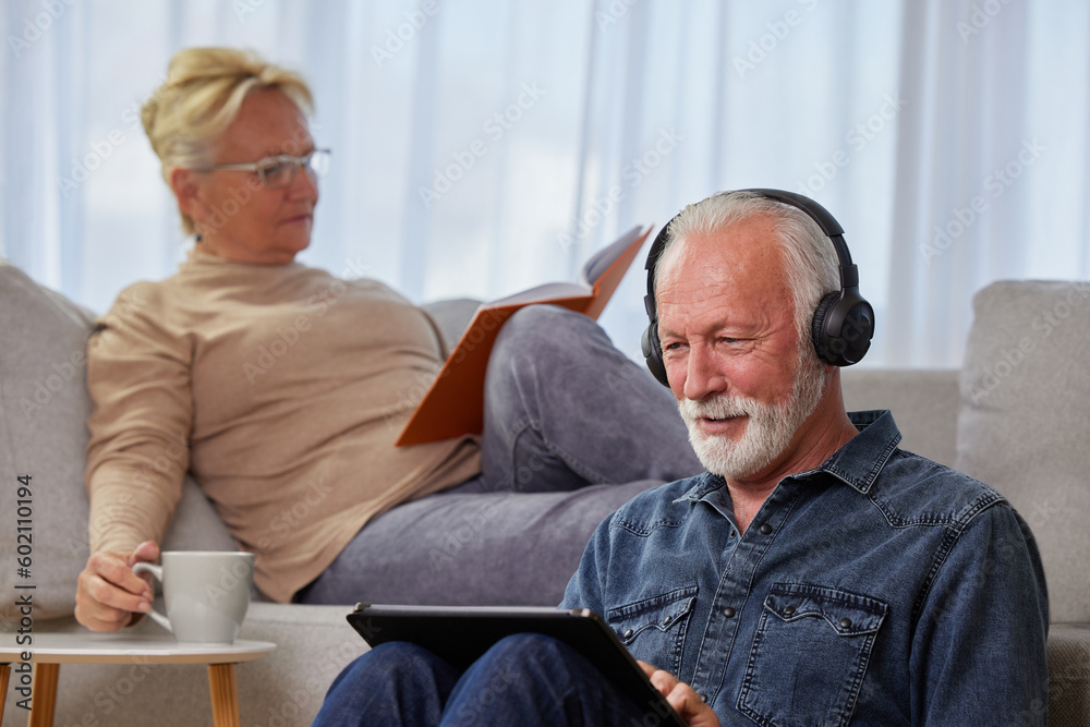 Senior couple spending leisure time at home. A woman reads a book, a man tablet pc and headphones listening to music, podcast or watching a movie while sitting on the floor. Happy retirement concept.