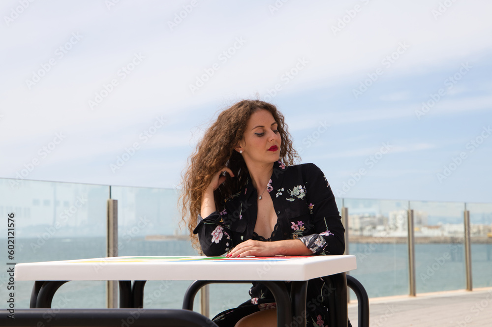 Beautiful blonde woman with curly hair and blue eyes is sitting on the promenade. In the background you can see the sea. The woman looks at camera and makes different expressions, happy, sad. Beautiful blonde woman with curly hair and blue eyes is sitting on the promenade. In the background you can see the sea. The woman looks at camera and makes different expressions, happy, sad.