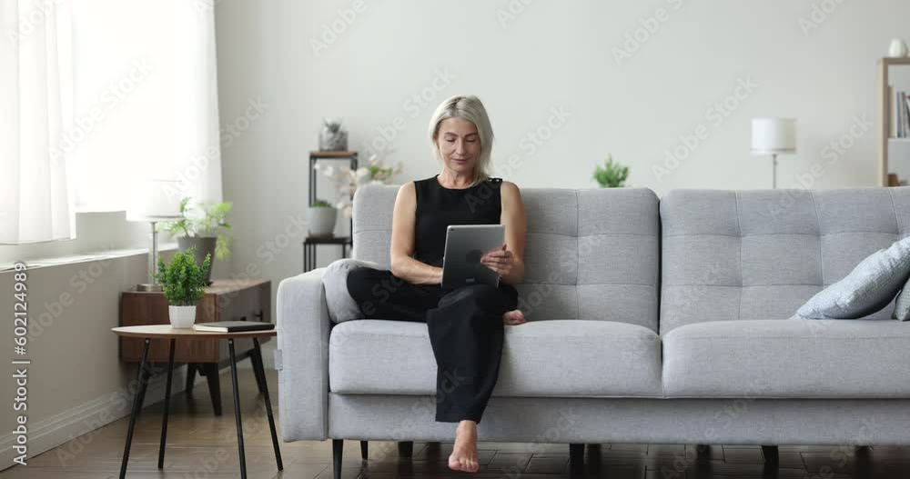 Focused mature woman sitting on home couch, typing on tablet, using online application, Internet media service, smiling, laughing, getting happy. Freelancer with gadget working from home Focused mature woman sitting on home couch, typing on tablet, using online application, Internet media service, smiling, laughing, getting happy. Freelancer with gadget working from home