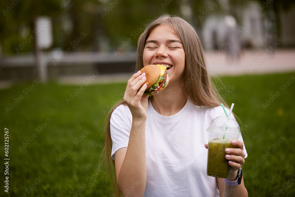 a young girl takes out a burger from a paper bag sitting on the green grass, the concept of food delivery, street food a young girl takes out a burger from a paper bag sitting on the green grass, the concept of food delivery, street food