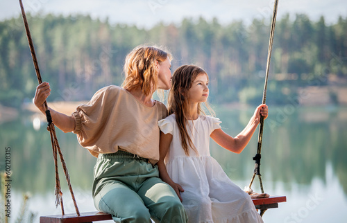Mom swings with her daughter on a rope swing against the backdrop of a forest lake reflecting the sky. Family concept photo