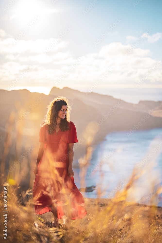 Front view of a smiling woman in red dress standing on a rock on a volcanic island in the Atlantic Ocean enjoying the morning atmosphere. São Lourenço, Madeira Island, Portugal, Europe.