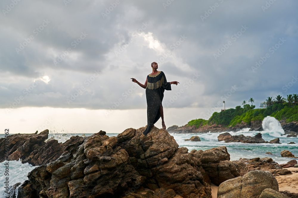 Gender fluid black person poses gracefully standing on rocks in ocean . Androgynous ethnic fashion model in posh dress and jewellery on rocky beach by storm. Pride month Gender fluid black person poses gracefully standing on rocks in ocean . Androgynous ethnic fashion model in posh dress and jewellery on rocky beach by storm. Pride month