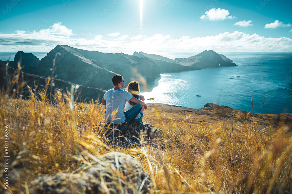 Back view of a tourist couple sitting on a rock, enjoying the view on coastal landscape of Madeira Island in the Atlantic Ocean in the morning. São Lourenço, Madeira Island, Portugal, Europe.