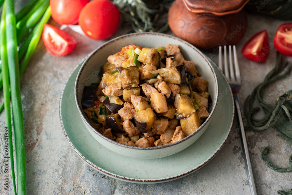 A hearty lunch for the family. A dish for dinner. Eggplant stewed with pork, spices and vegetables in a beautiful plate on a gray background. Close-up. A hearty lunch for the family. A dish for dinner. Eggplant stewed with pork, spices and vegetables in a beautiful plate on a gray background. Close-up.