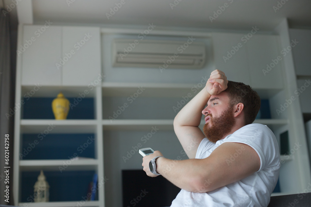 Young man with air conditioner remote control using air conditioner at home, guy is cooling off during hot weather at summer, suffering from heat, high temperature. Broken AC 