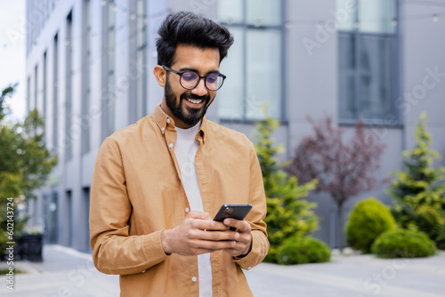 Successful Indian young businessman outside office building walking in daytime, man holding phone in hands, businessman dialing, browsing online pages, programmer engineer in glasses. photo