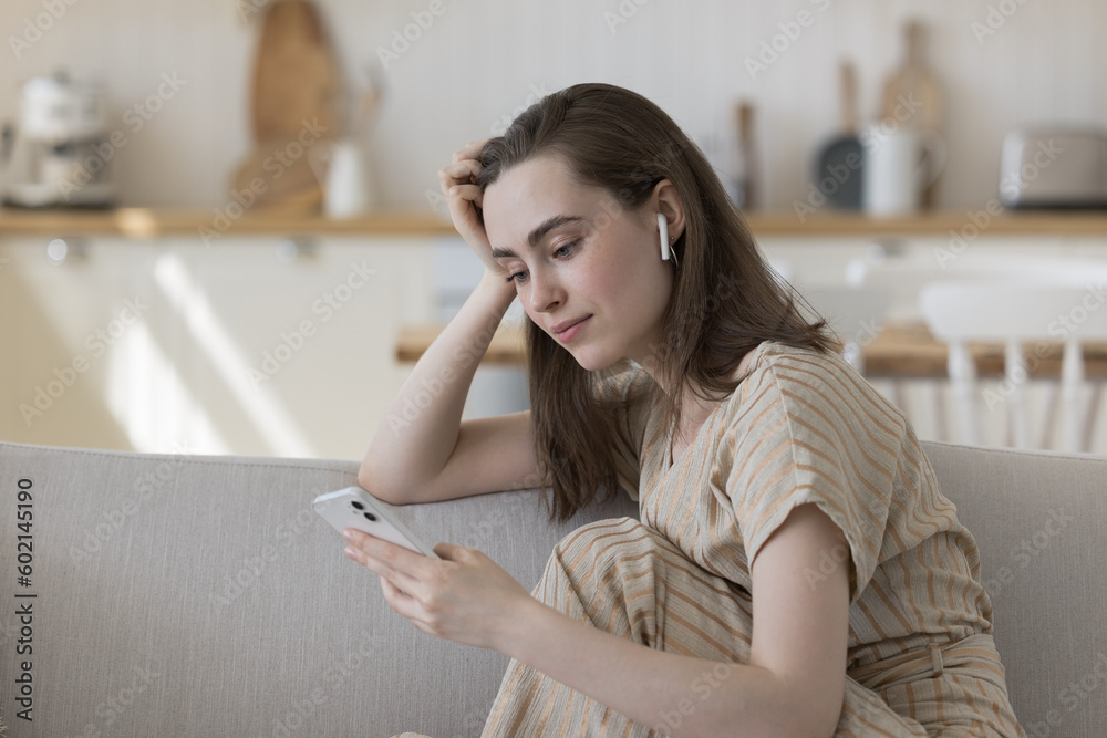 Calm relaxed adult girl using smartphone and small wireless earphones in ears, resting on home couch, enjoying leisure, online Internet communication, listening to music, turning player on