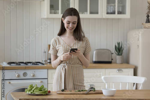 Positive adult girl browsing Internet on smartphone in kitchen  consulting cook blog  reading online recipe  typing message  preparing salad from fresh vegetables  taking picture of ingredients