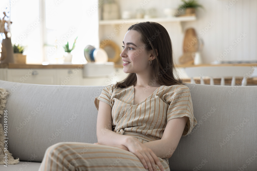 Happy dreamy beautiful young woman sitting on comfortable sofa, relaxing, enjoying comfort, leisure, break, looking away, laughing, smiling at good thoughts, thinking over plans Happy dreamy beautiful young woman sitting on comfortable sofa, relaxing, enjoying comfort, leisure, break, looking away, laughing, smiling at good thoughts, thinking over plans