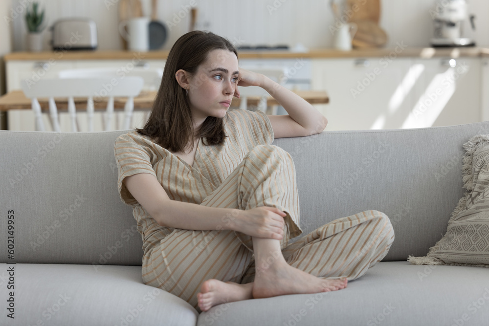 Frustrated depressed adult girl sitting on home sofa, looking away, thinking over troubles, health problems, suffering from depression, apathy, loneliness, coping with loss, emotional crisis Frustrated depressed adult girl sitting on home sofa, looking away, thinking over troubles, health problems, suffering from depression, apathy, loneliness, coping with loss, emotional crisis