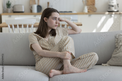 Frustrated depressed adult girl sitting on home sofa, looking away, thinking over troubles, health problems, suffering from depression, apathy, loneliness, coping with loss, emotional crisis
