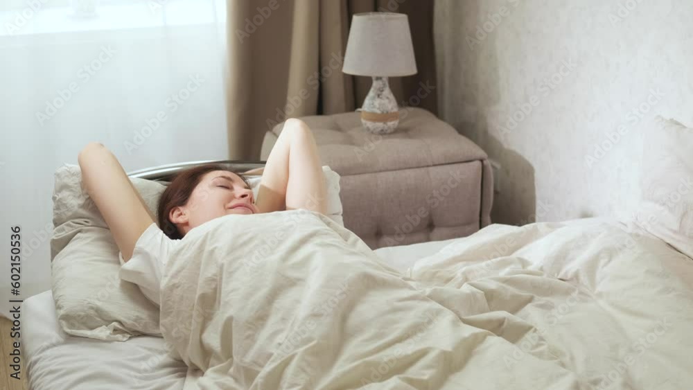 An attractive young woman wakes up in the bedroom and stretches in bed in the early sunny morning, smiles and sits down An attractive young woman wakes up in the bedroom and stretches in bed in the early sunny morning, smiles and sits down