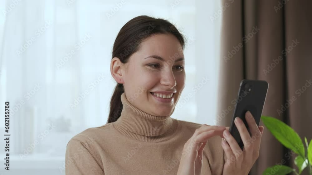 Attractive young adult Caucasian woman brunette in beige sweatshirt uses the phone in the room and smiles Attractive young adult Caucasian woman brunette in beige sweatshirt uses the phone in the room and smiles