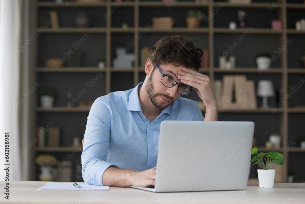 Tired sad freelancer man getting problems with laptop, software error, feeling stress, exhaustion, headache, touching head at home work table, looking at screen with doubt Tired sad freelancer man getting problems with laptop, software error, feeling stress, exhaustion, headache, touching head at home work table, looking at screen with doubt