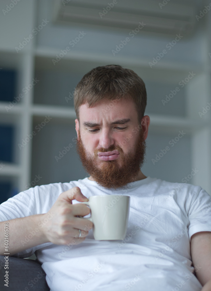 Unsatisfied, sad, upset man with mug, man does not want to take the medicine, beverage man is sitting in the living room, at home Unsatisfied, sad, upset man with mug, man does not want to take the medicine, beverage man is sitting in the living room, at home