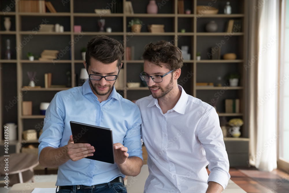 Positive successful family business startup owners talking, discussing online project. Young twin brothers in glasses and office shirts using tablet at home, cooperating on Internet technology product