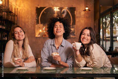 Three women friends pose smiling for photo while spending free time having a drink at cafe shop. Cheerful and happy girls looking at camera having fun together. People gathered inside drinking coffee.