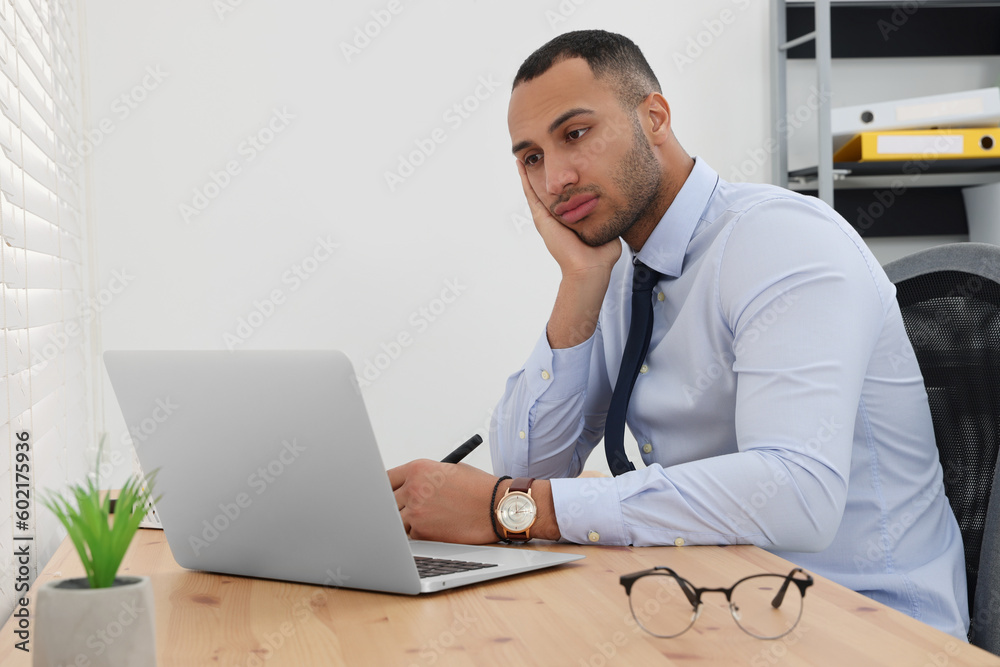 Puzzled young intern using laptop at table in modern office. First work day Puzzled young intern using laptop at table in modern office. First work day