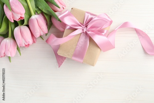 Beautiful gift box with bow and pink tulip flowers on white wooden background, flat lay. Space for text