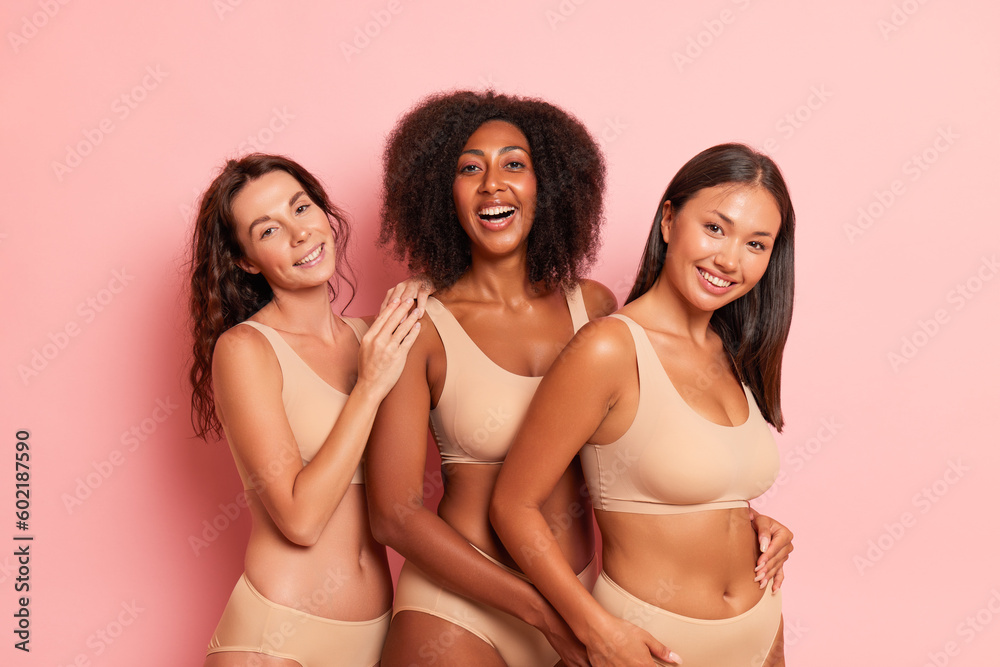 Three very happy girlfriends wearing beige tops and panties pose inside, black-skined girl in the middle her friends are on both sides, comfort underwear concept, copy space - obrazy, fototapety, plakaty 