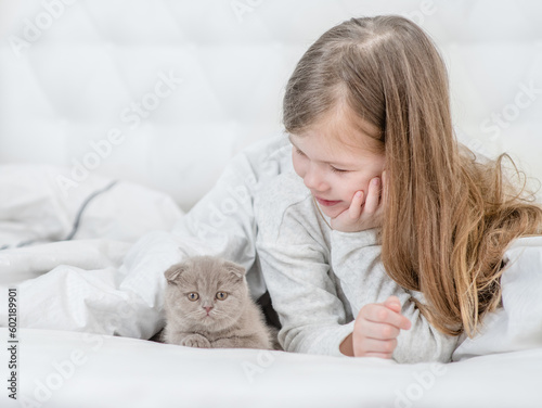 Happy little girl and tiny kitten lying together on the bed at home