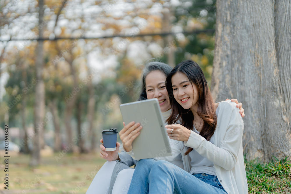 Fototapeta premium Grown daughter with aging mother expressing love and looking at tablet computer under tree in public park.