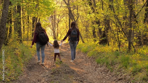 Happy family life concept. young women walk through forest with small child with backpacks their backs. walk woods summer weekend sunset. little girl holding hands parents walking along path summer