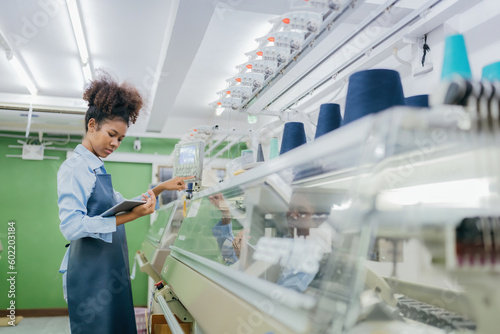 american female designer In hand, there is a tablet to see customer orders for production. And she is using the control panel of a sewing machine to design clothing in a large weaving factory.