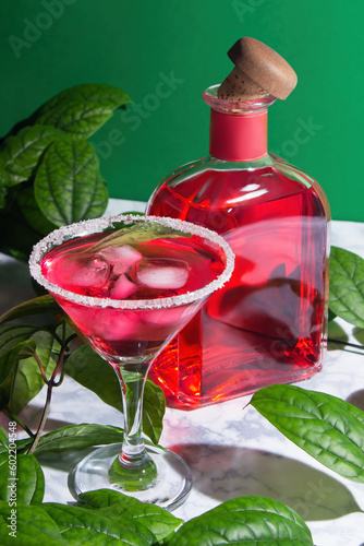 Red cocktail with ice in a margarita glass with a sugar rim, next to it is red alcohol in a bottle with a wooden cork.  Surrounded by green leaves of the plant, on a green background. photo