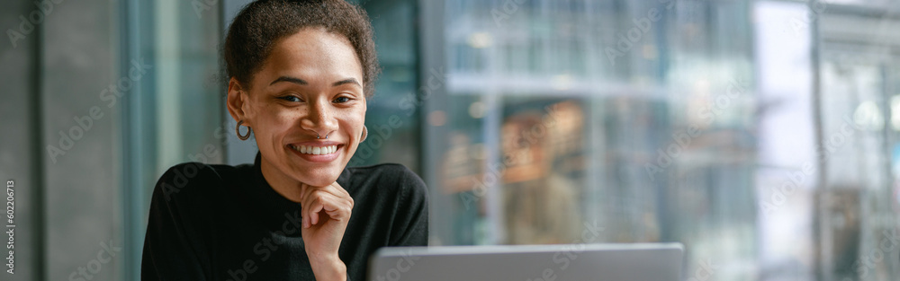 Smiling female student studying on laptop while sitting in cafe and looking on camera