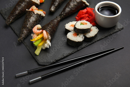 Slate board with tasty sushi cones, rolls, soy sauce, ginger and chopsticks on dark background, closeup