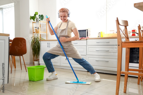 Young woman with headphones mopping floor in kitchen