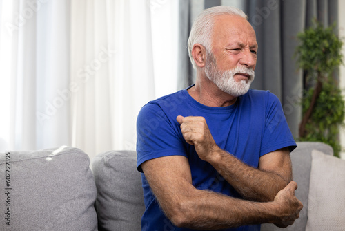 Elderly man holding his elbow, suffering from arthritis pain in hand, sitting on sofa at home