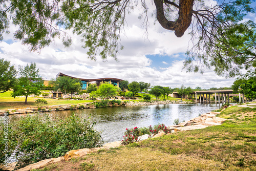 A visitor center on the other side of a tranquil river in San Angelo, Texas, surrounded by lush grass and trees with wispy clouds dotting a bright blue sky. photo