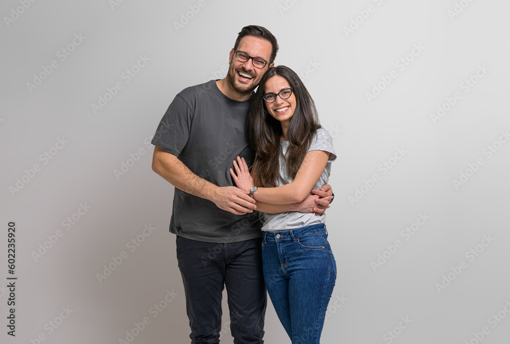 Portrait of loving young couple embracing and holding hands while posing ecstatically against background. Charming girlfriend and boyfriend wearing eyeglasses standing together and looking at camera - obrazy, fototapety, plakaty 