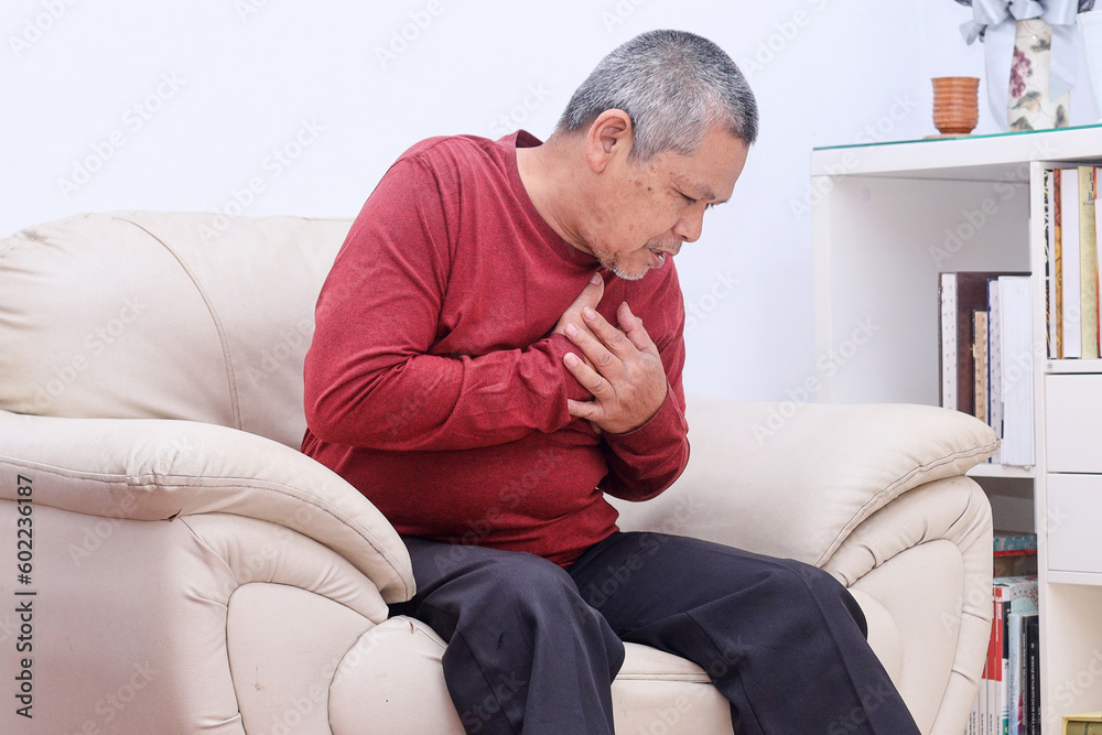 Elderly Asian male clutching his left chest from acute pain while sitting at the couch in the living room. 