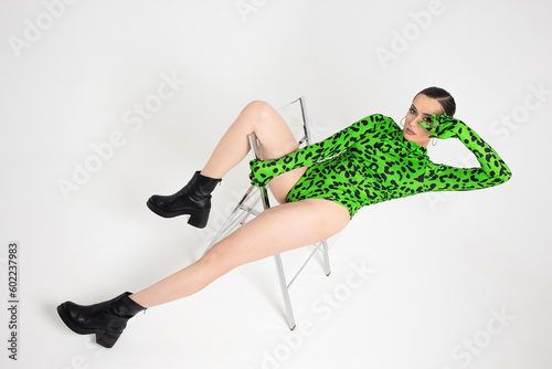 Stylish bright dancer in a bright green leopard print bodysuit, posing on a chair, studio photo on a white background. Plastic body, pretentious pose