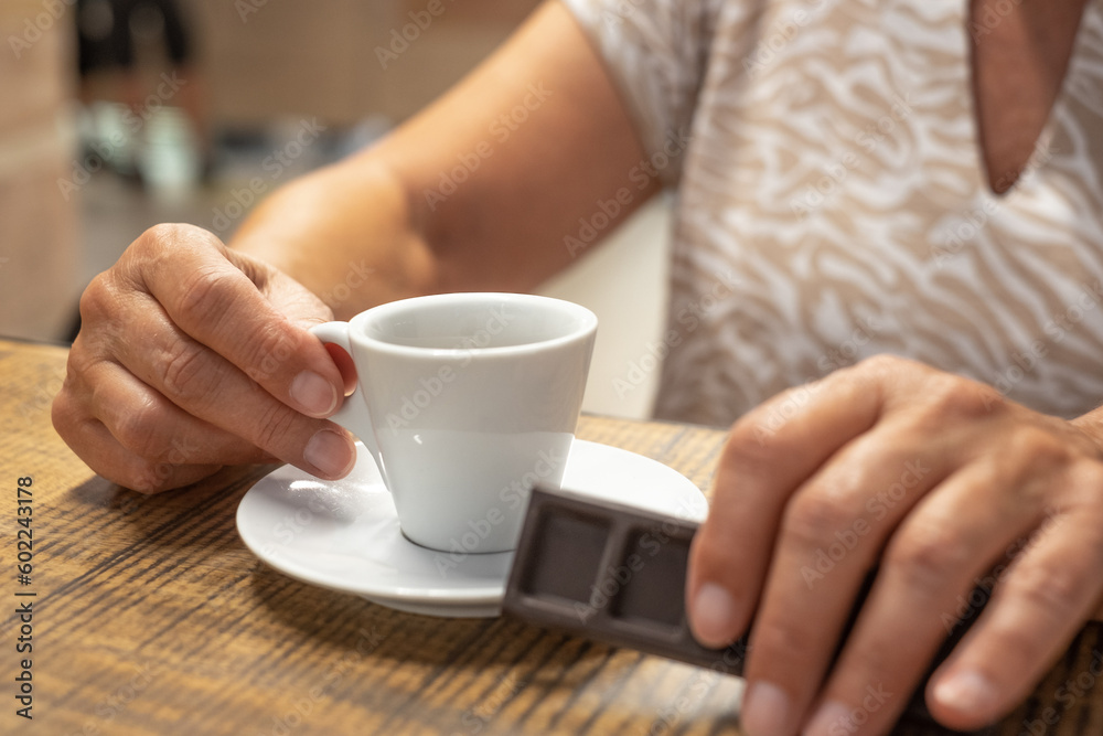 Old caucasian senior woman holding dark chocolate bar in hand while enjoying cup of espresso coffee - break time with hot aromatic drink